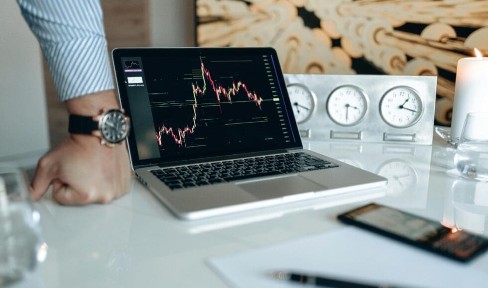 Laptop displaying a stock market graph on a desk with clocks and documents.